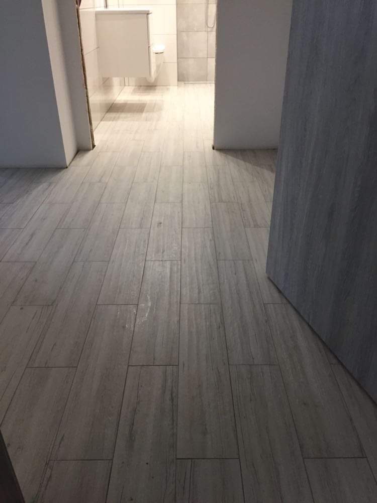 Ułożona podłużna, szara, matowa glazura imitująca drewno na podłodze w łazience z widoczną umywalką i kabiną prysznicową w tle.