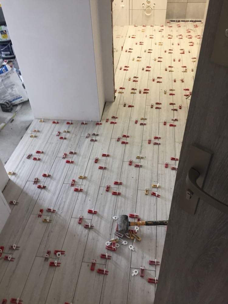 Układanie płytek podłogowych w łazience, widoczne krzyżyki dystansowe i gumowy młotek, płytki imitujące drewno