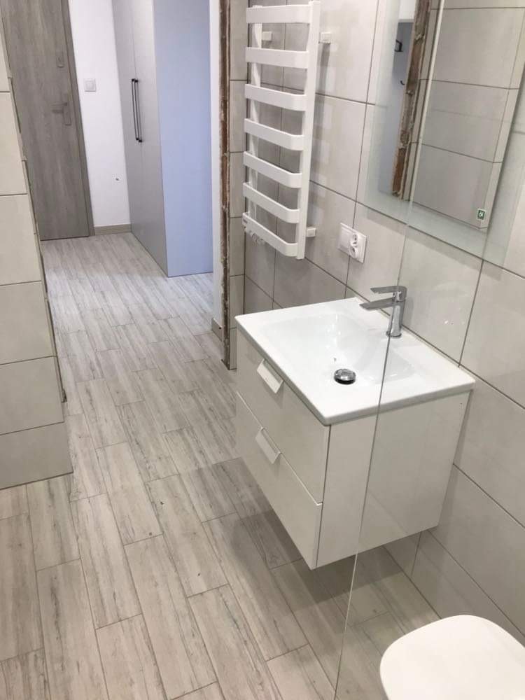 Nowoczesna łazienka z białymi płytkami imitującymi drewno, wisząca szafka z umywalką, grzejnik drabinkowy i częściowo widoczna kabina prysznicowa.