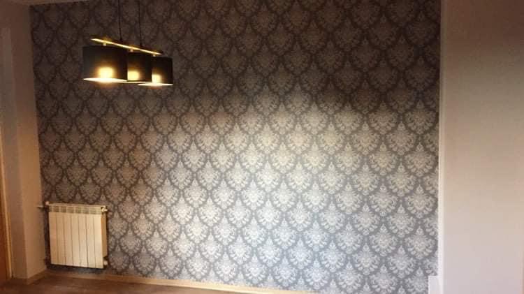 Ściana pokryta szarą tapetą w ornamentowy wzór, oświetlona wiszącą lampą z trzema czarnymi kloszami, widoczny biały grzejnik pod ścianą.