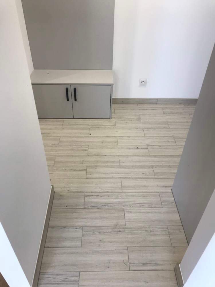 Wykończona podłoga z płytek imitujących drewno w jasnym odcieniu, zabudowa meblowa pod sufit w kolorze szarym, widoczny fragment gniazdka elektrycznego na białej ścianie.