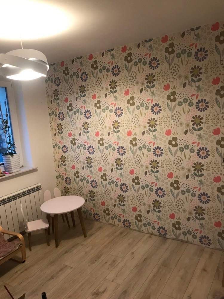 Pokój dziecięcy po odświeżeniu: ściana w kwieciste tapety, drewniana podłoga, okrągły stolik i krzesełka w pastelowych odcieniach, nowoczesna lampa sufitowa.