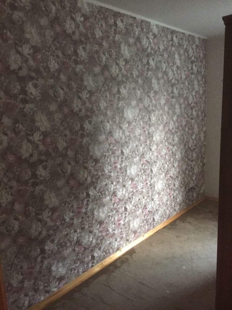 Ściana pokoju pokryta tapetą w kwiaty w odcieniach szarości i różu, widoczna listwa przypodłogowa i fragment podłogi.