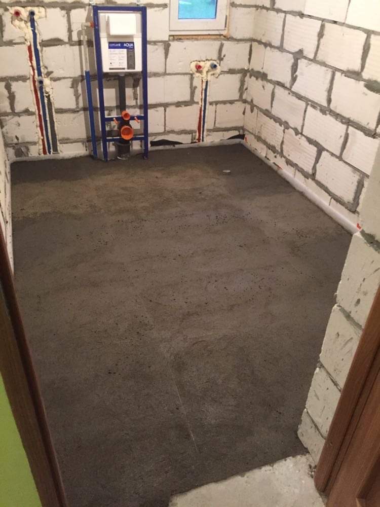 Surowy stan łazienki przed wykończeniem: wylewka betonowa na podłodze, ściany z białego bloczka, instalacja podtynkowego WC z niebieską ramą montażową i widocznymi rurami doprowadzającymi wodę.