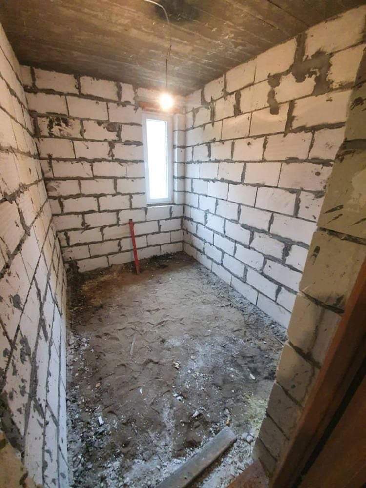 Surowy stan łazienki w trakcie budowy: ściany z białych bloczków, surowy betonowy sufit, małe okno, ziemia na podłodze, widoczna instalacja elektryczna.