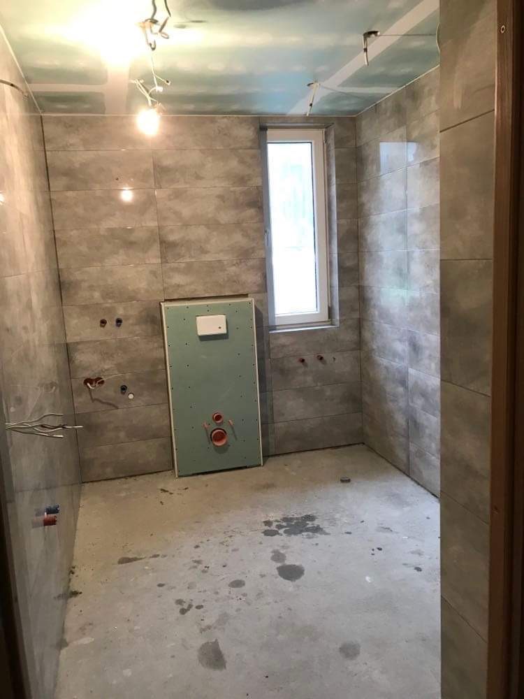 Wnętrze łazienki w trakcie prac: szare płytki na ścianach, surowy beton na podłodze z plamami, zamontowane okno, oraz zabudowa stelaża WC z widocznymi otworami na instalacje.