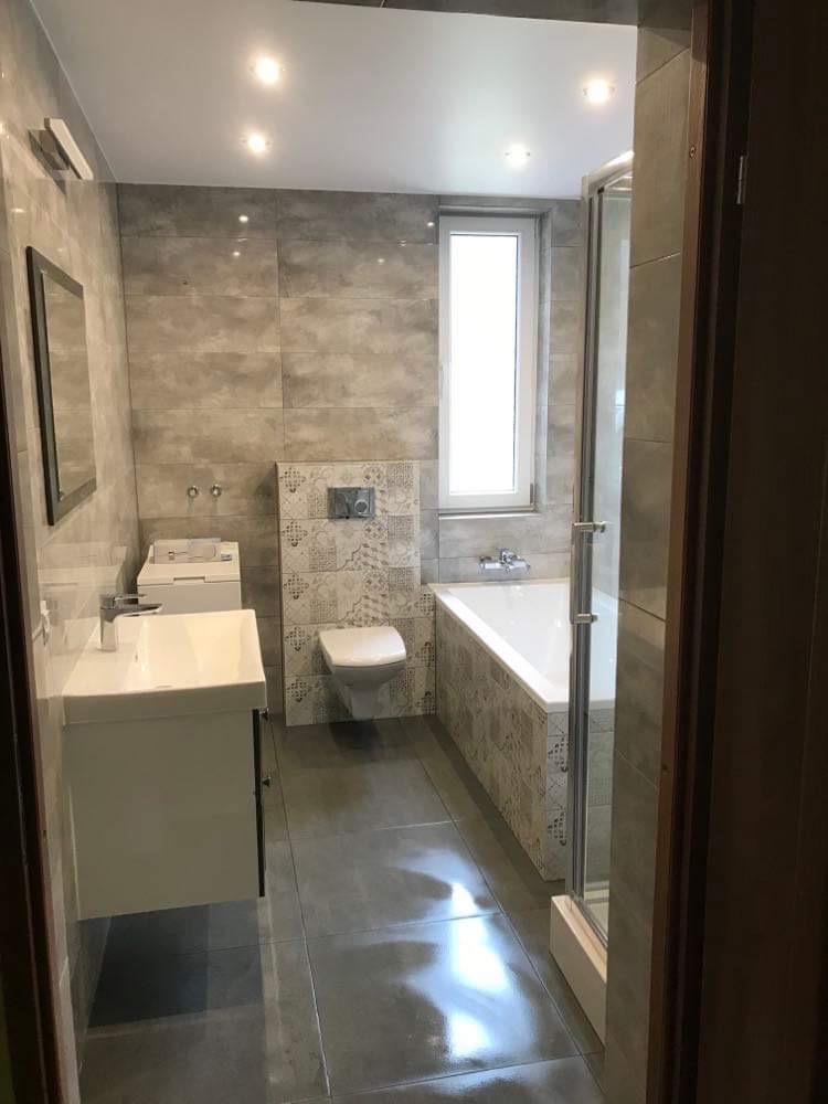 Wnętrze wykończonej łazienki z szarymi płytkami imitującymi kamień, białą armaturą, kabiną prysznicową z przezroczystego szkła i oświetleniem punktowym.