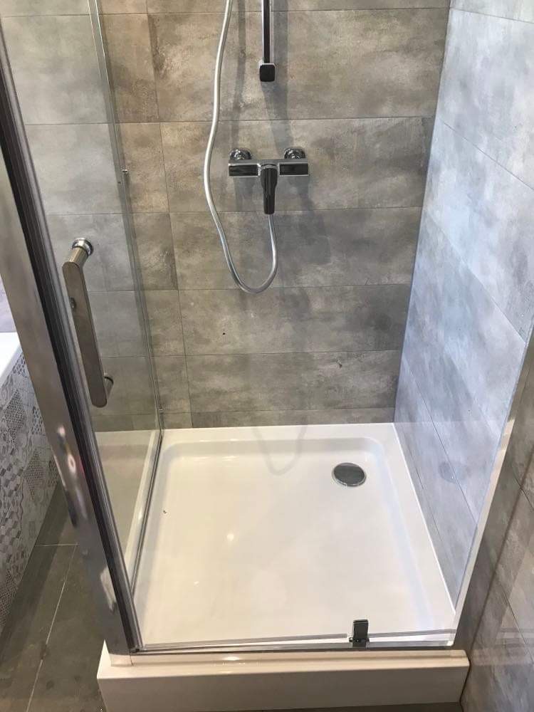 Nowoczesny brodzik z kabiną prysznicową, szare płytki imitujące beton na ścianach, chromowana bateria i deszczownica, widoczny fragment wanny z ozdobnymi płytkami.