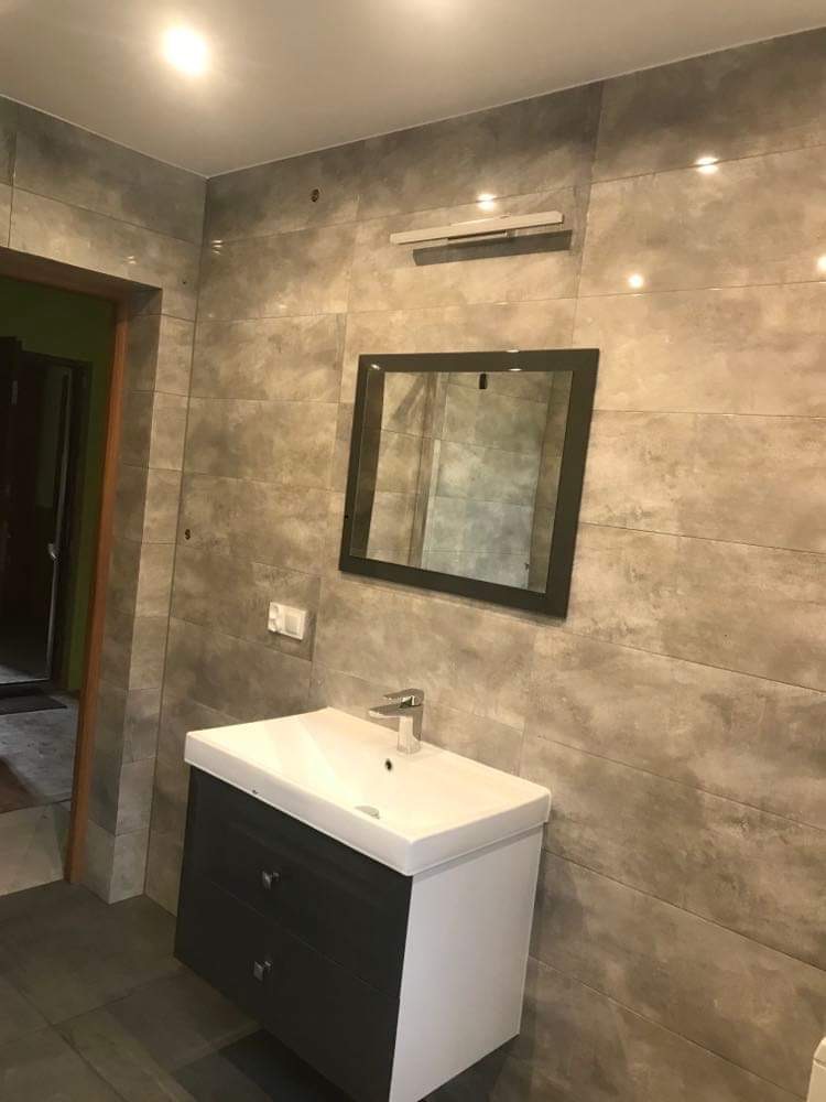 Wykończona łazienka z szarymi płytkami imitującymi beton, wiszącą szafką z umywalką i lustrem w czarnej ramie, oświetlona lampą nad lustrem.