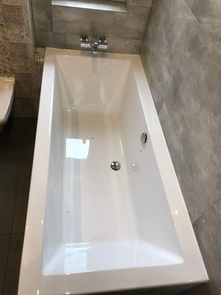 Nowa, biała wanna z chromowaną armaturą zamontowana w łazience wykończonej szarą, wielkoformatową płytką imitującą beton. Widoczny odpływ i przelew wanny.