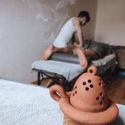 Masażysta wykonuje masaż plec&oacute;w klientowi na stole do masażu; w pierwszym planie ceramiczny dyfuzor zapachowy z widocznym dymem.