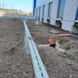 Długi, betonowy kanał odwadniający z kratką szczelinową, wkopany w ziemię przed budynkiem przemysłowym z rampami załadunkowymi, rury odpływowe w kolorze terakoty, widoczne elementy kanalizacji...