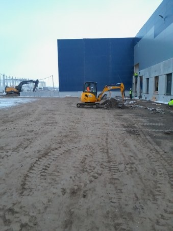 Żółta mini koparka JCB podczas prac ziemnych przy budowie hali, widoczne ślady opon na błotnistym podłożu i operator w kabinie, w tle druga koparka i konstrukcja stalowa budynku.