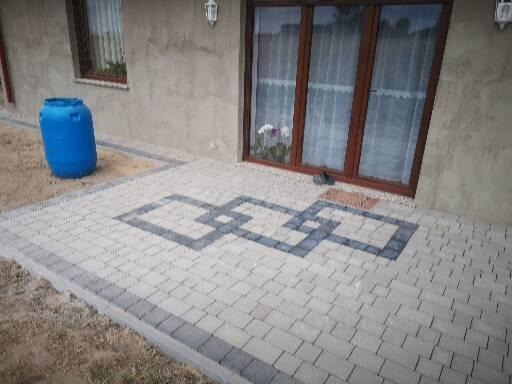 Ułożona kostka brukowa przed domem z wzorem geometrycznym w szarych odcieniach, widoczny niebieski pojemnik na deszczówkę i brązowe okno z białymi firankami.