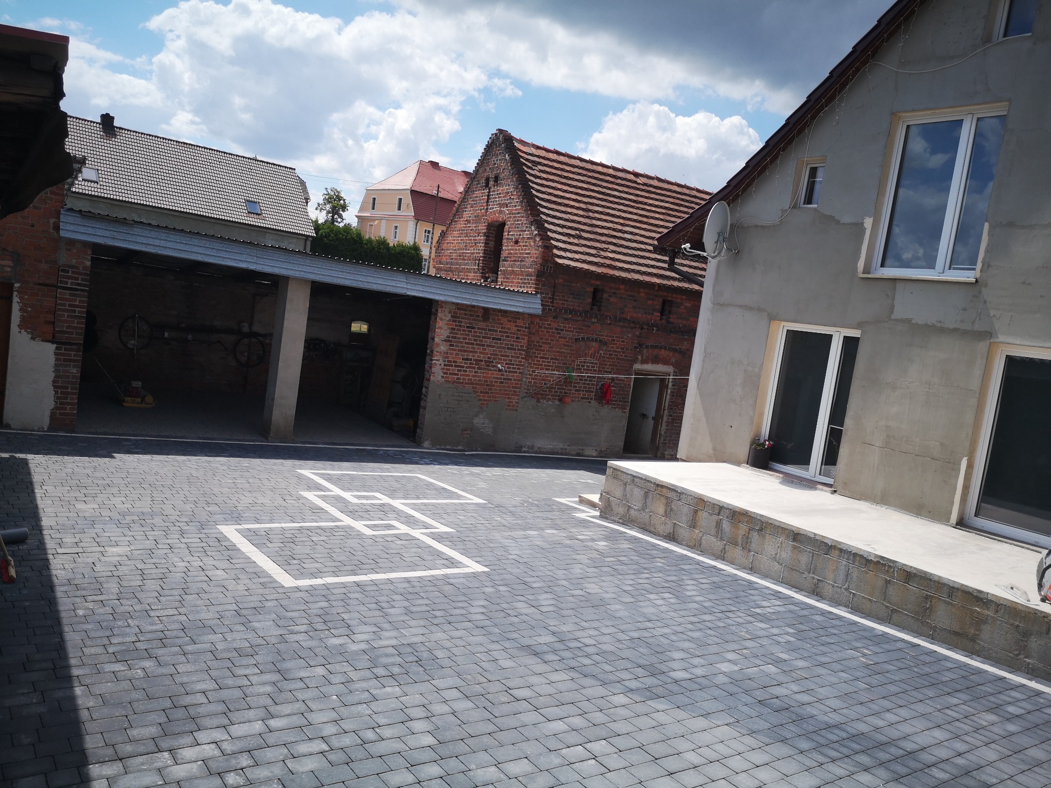 Ułożona szara kostka brukowa tworząca podjazd z wyznaczonymi białymi liniami miejsc parkingowych, na tle budynków mieszkalnych.