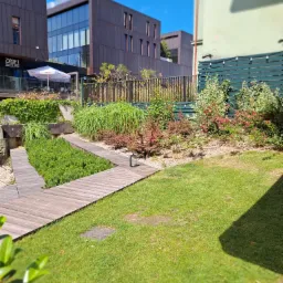 Nowoczesny ogród z drewnianą ścieżką, ozdobnymi roślinami i trawą, na tle budynków o architekturze industrialnej.