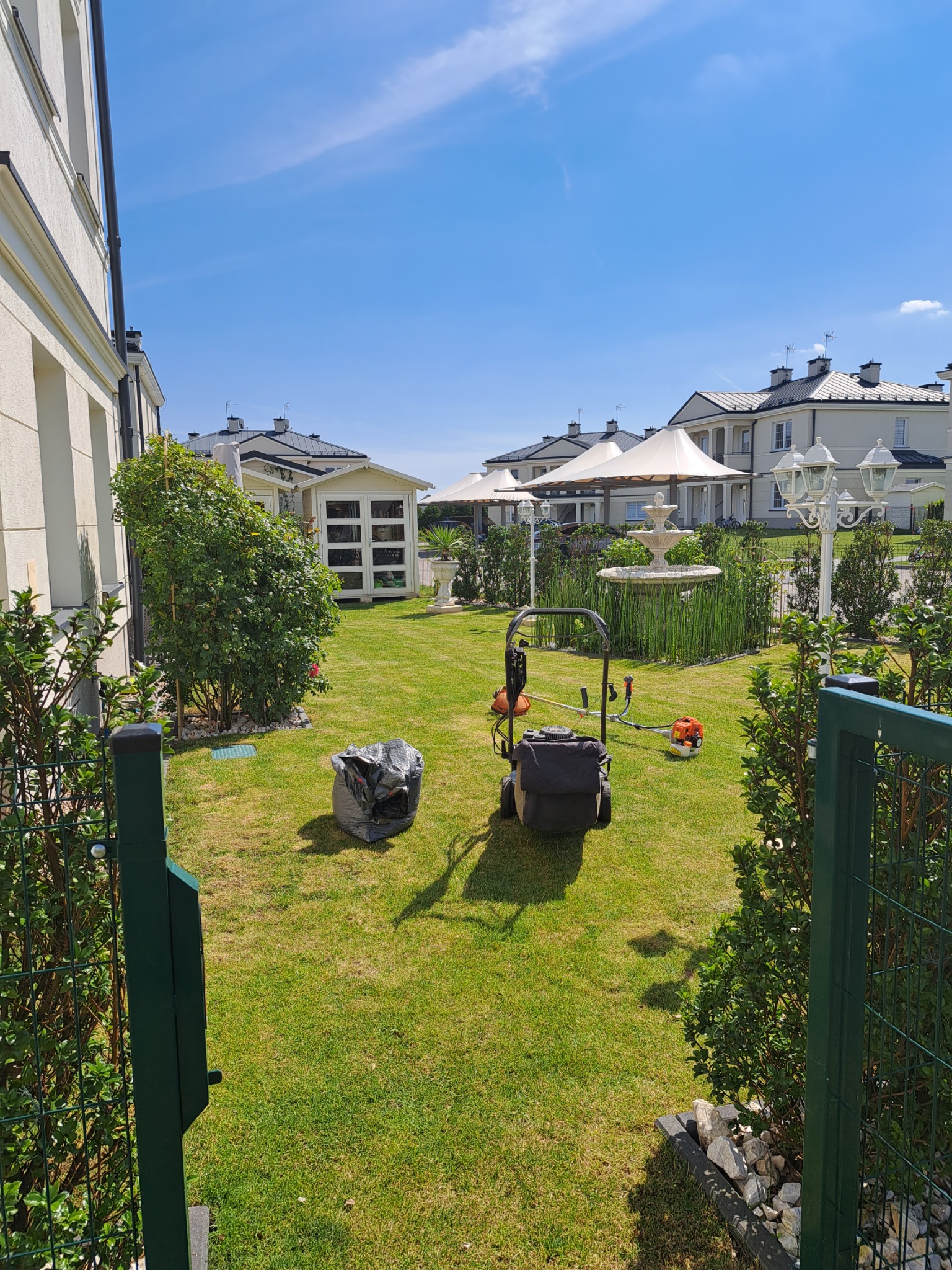 Świeżo skoszony trawnik w zadbanym ogrodzie z fontanną, narzędziami ogrodniczymi i workiem na odpady, widok zza zielonej bramy.