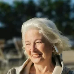 Uśmiechnięta starsza kobieta z siwymi włosami i słuchawką bezprzewodową w uchu, ubrana w bluzę w kratę, na tle rozmytego krajobrazu z drzewami i błękitnym niebem.