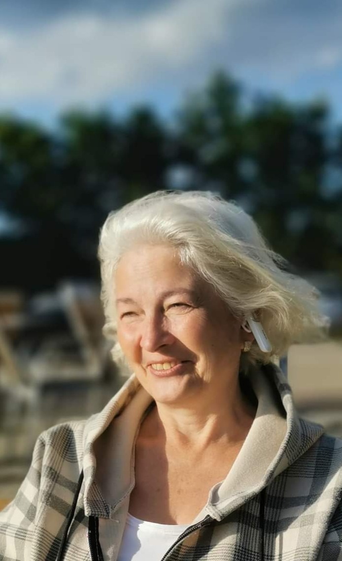 Uśmiechnięta starsza kobieta z siwymi włosami i słuchawką bezprzewodową w uchu, ubrana w bluzę w kratę, na tle rozmytego krajobrazu z drzewami i błękitnym niebem.