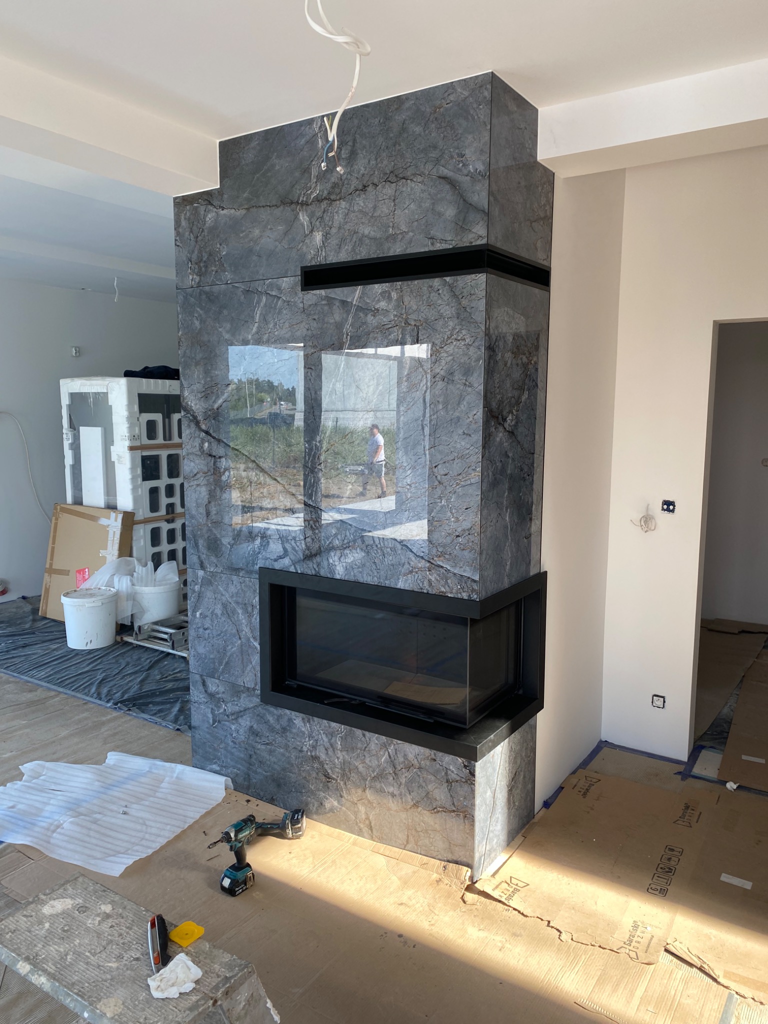 Obudowa kominka z szarego marmuru z czarną ramą, w trakcie montażu, widoczne narzędzia i zabezpieczenia podłogi.