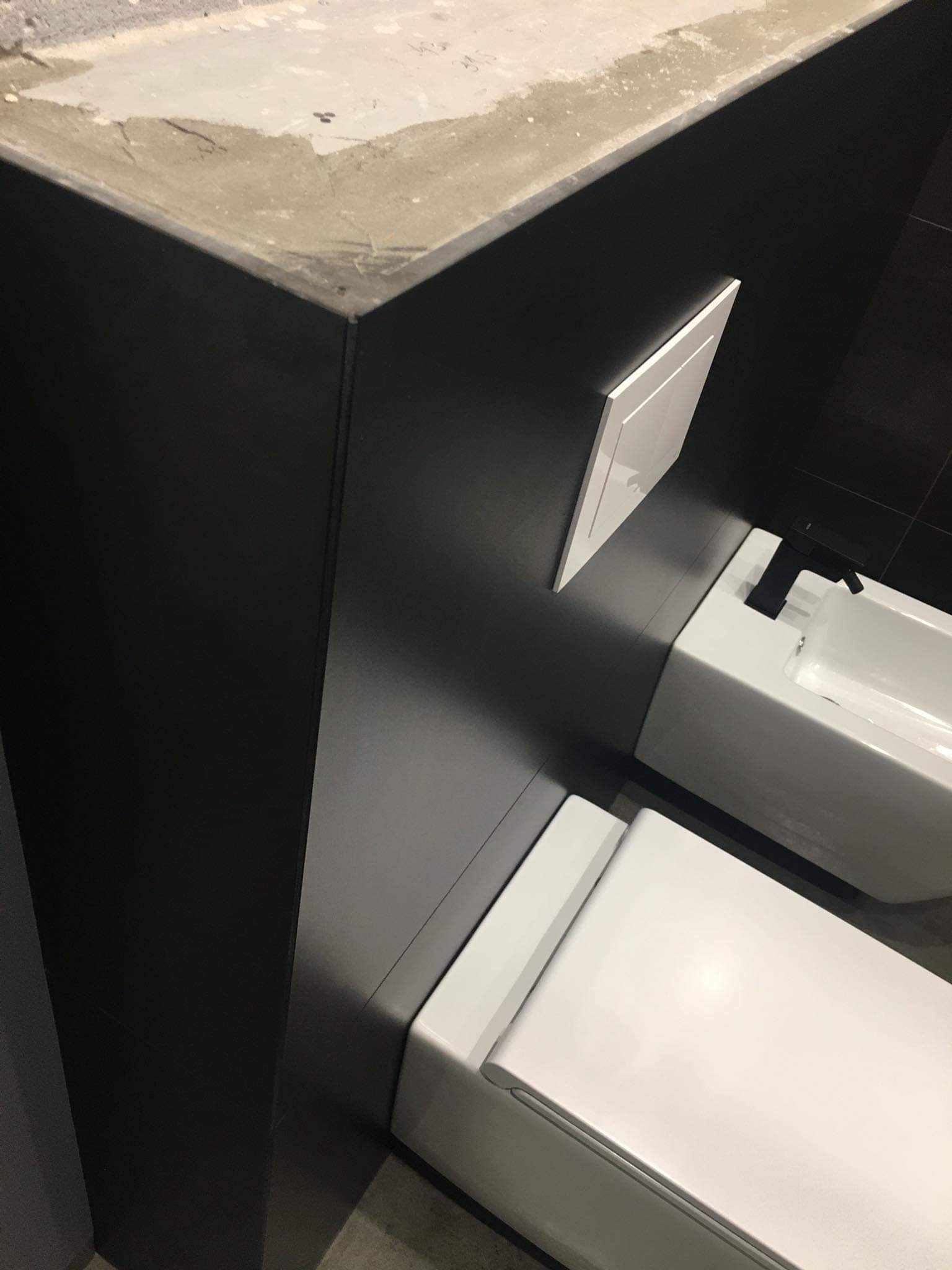 Nowoczesna łazienka z czarnymi płytkami, białą armaturą i podtynkową spłuczką, widok z góry na surowy betonowy element wykończeniowy.