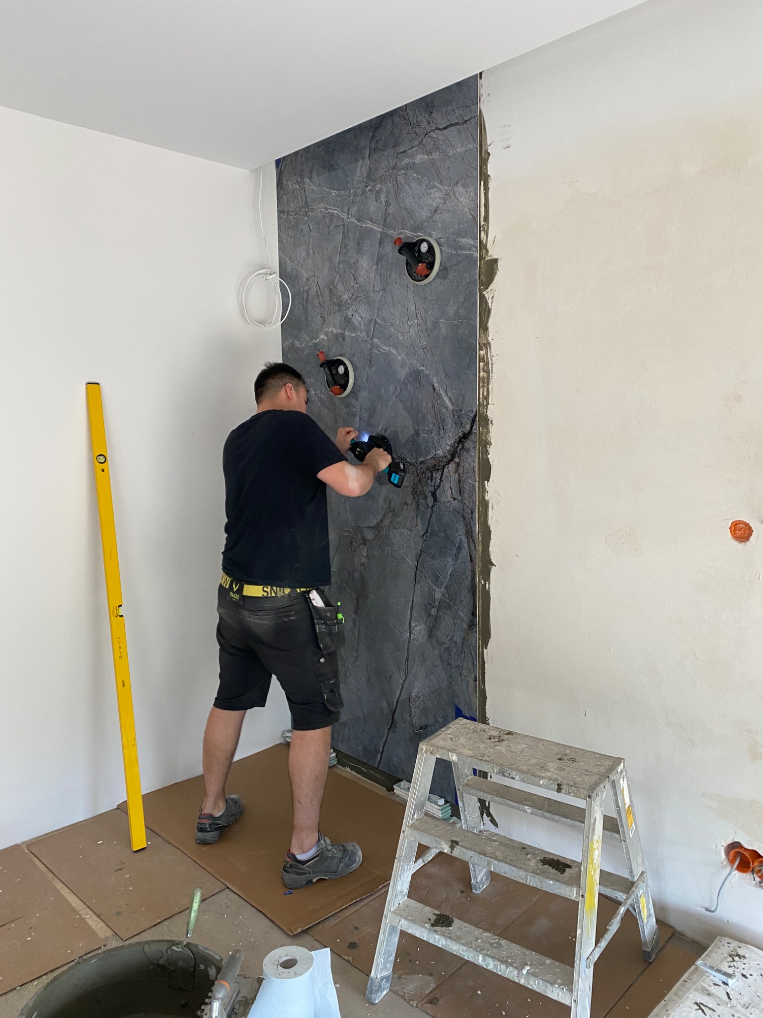 Mężczyzna w czarnej koszulce wierci otwory w szarej płycie imitującej kamień, zamontowanej na ścianie w trakcie prac wykończeniowych w łazience; widoczna poziomica, drabina i wiadro z zaprawą.
