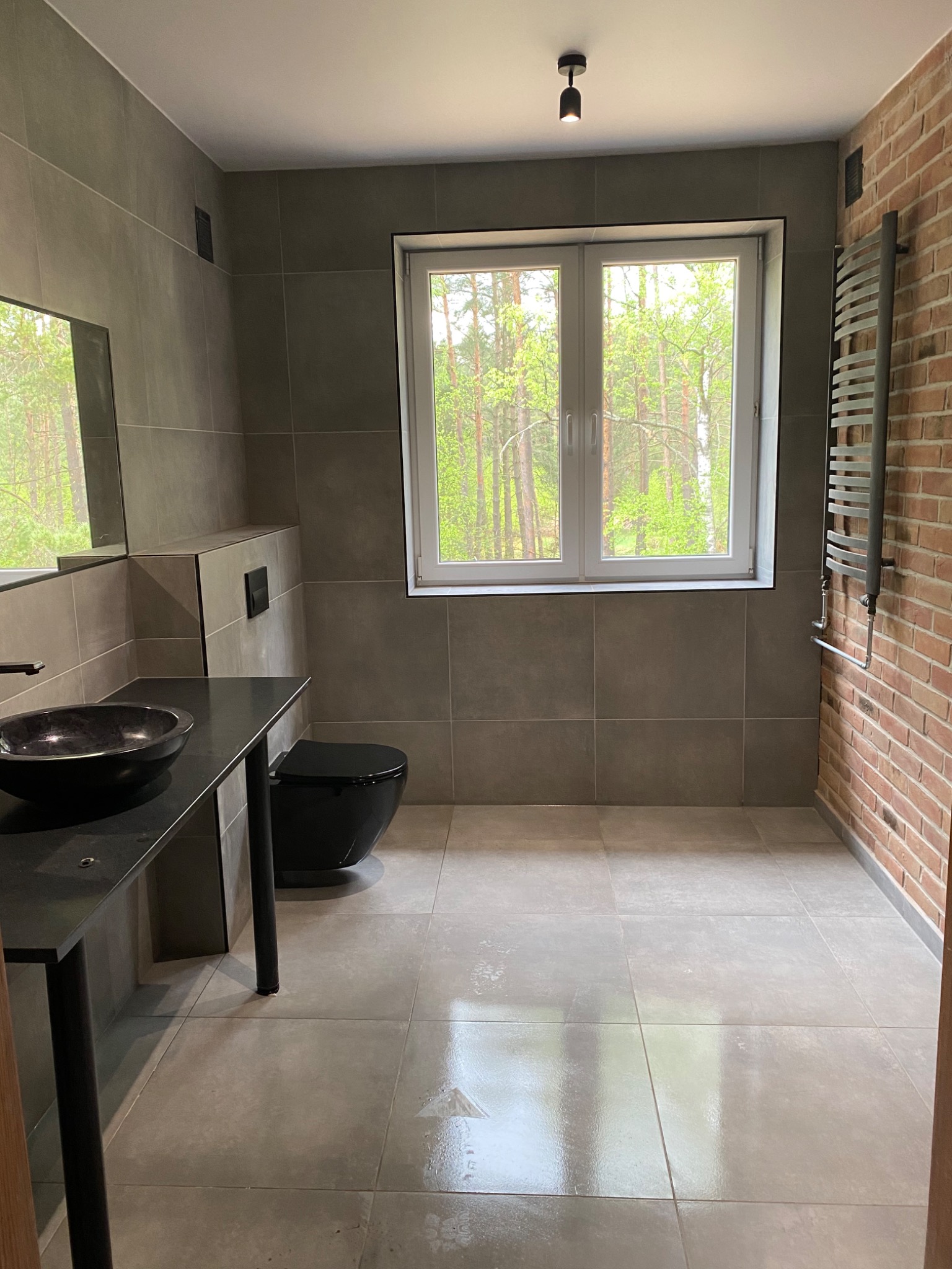 Nowoczesna łazienka z szarymi płytkami, czarną umywalką w kształcie misy, wiszącą toaletą i ceglaną ścianą z grzejnikiem drabinkowym, widok na las za oknem.