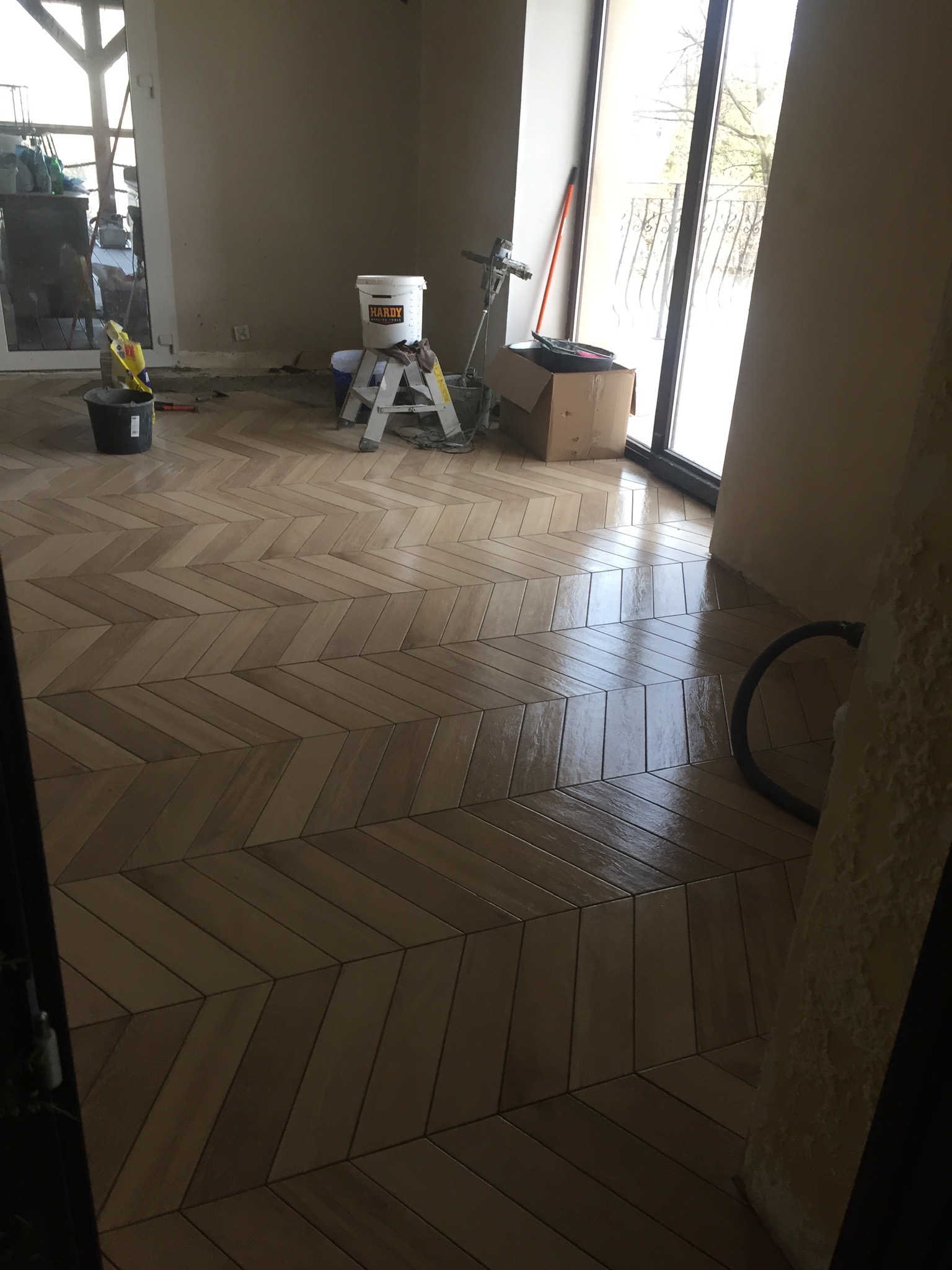 Ułożona w jodełkę podłoga z płytek imitujących drewno, widoczne narzędzia i materiały budowlane w tle, wysokie okno balkonowe z prawej strony.