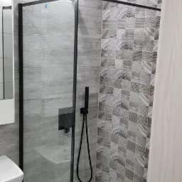Nowoczesna kabina prysznicowa z czarną armaturą i deszczownicą, szare płytki imitujące kamień oraz dekoracyjne płytki mozaikowe w odcieniach szarości i beżu.