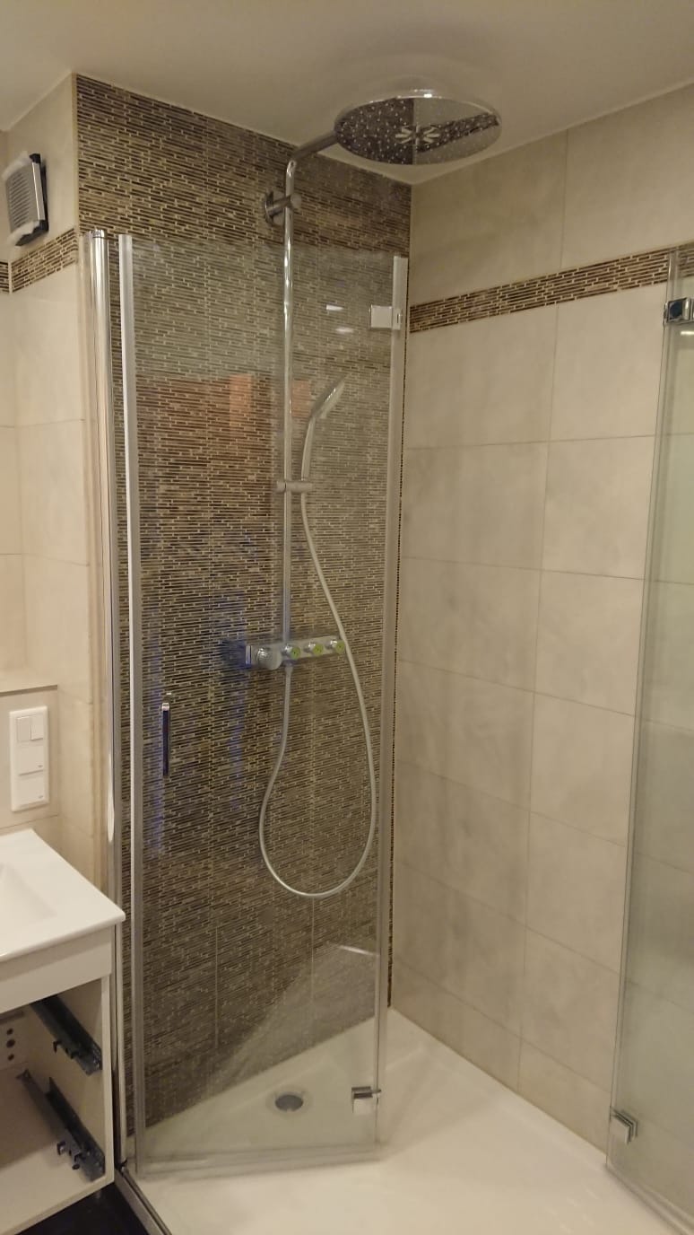 Nowoczesna kabina prysznicowa z deszczownicą i baterią termostatyczną, wykończona mozaiką w odcieniach brązu i beżu, widok z boku, z elementami szafki umywalkowej po lewej stronie.