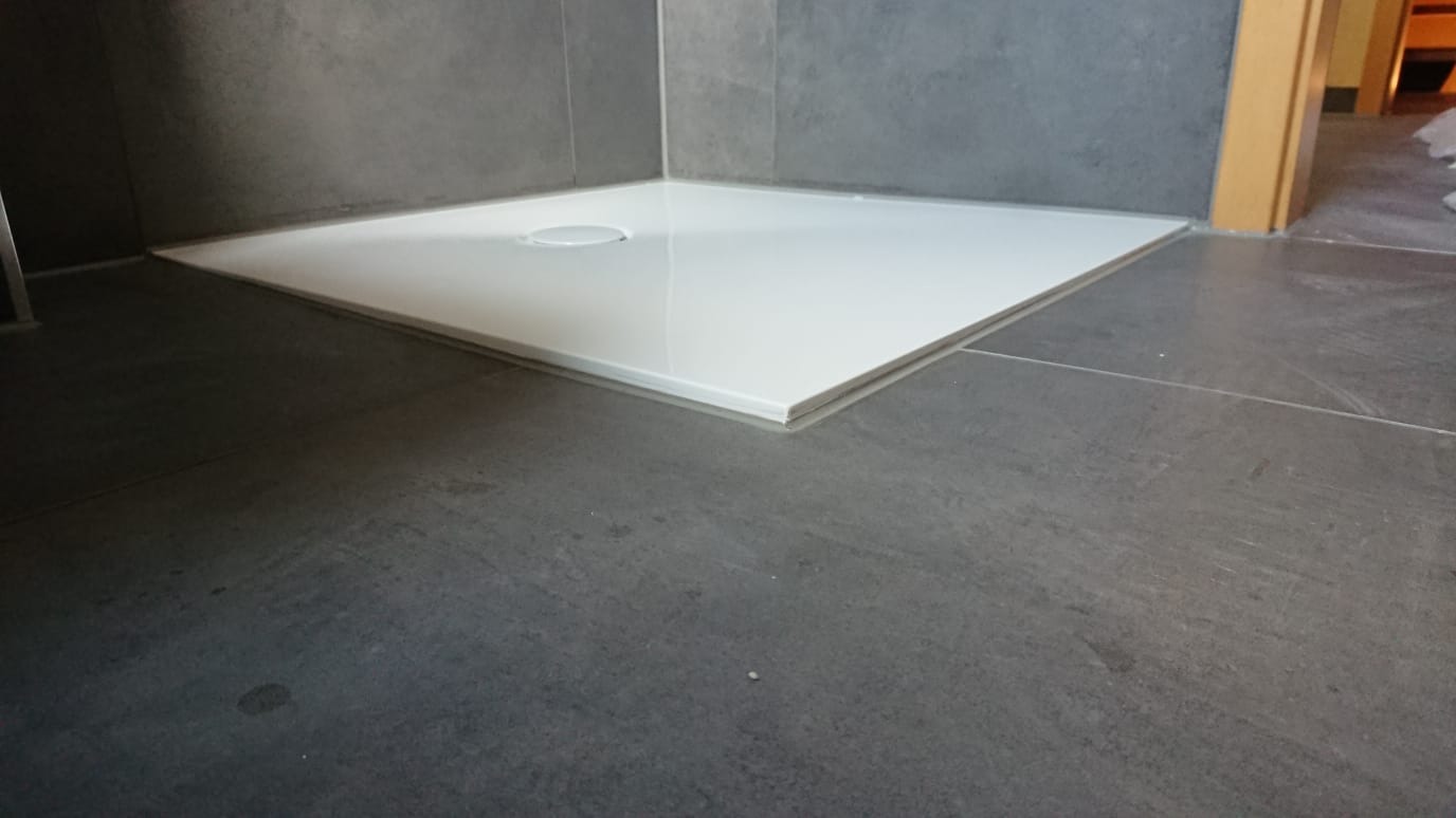 Biały, kwadratowy brodzik z odpływem w rogu, osadzony w szarej, płytkowej podłodze i ścianach łazienki, z widocznym fragmentem futryny drzwiowej w tle.