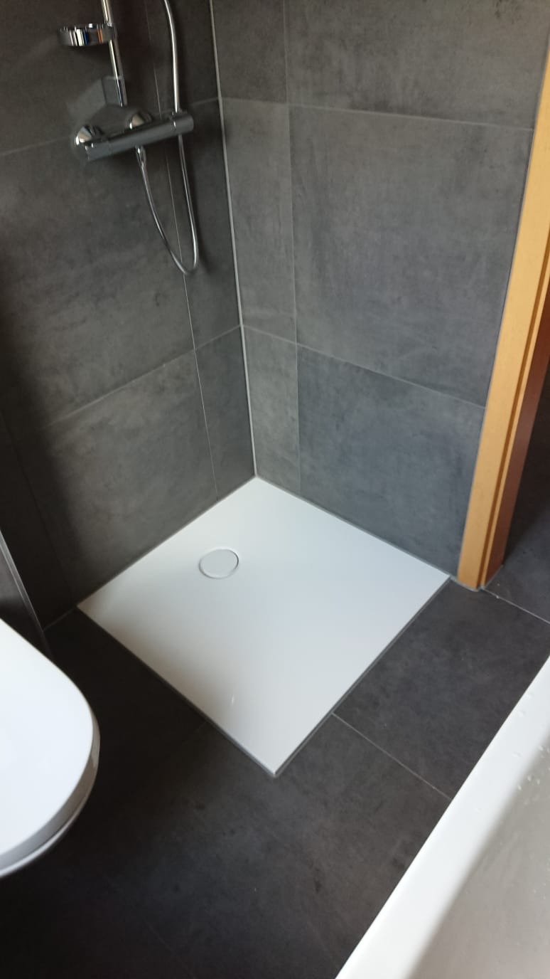 Nowoczesna kabina prysznicowa z białym brodzikiem i szarymi płytkami imitującymi beton, widoczna bateria prysznicowa i fragment toalety.