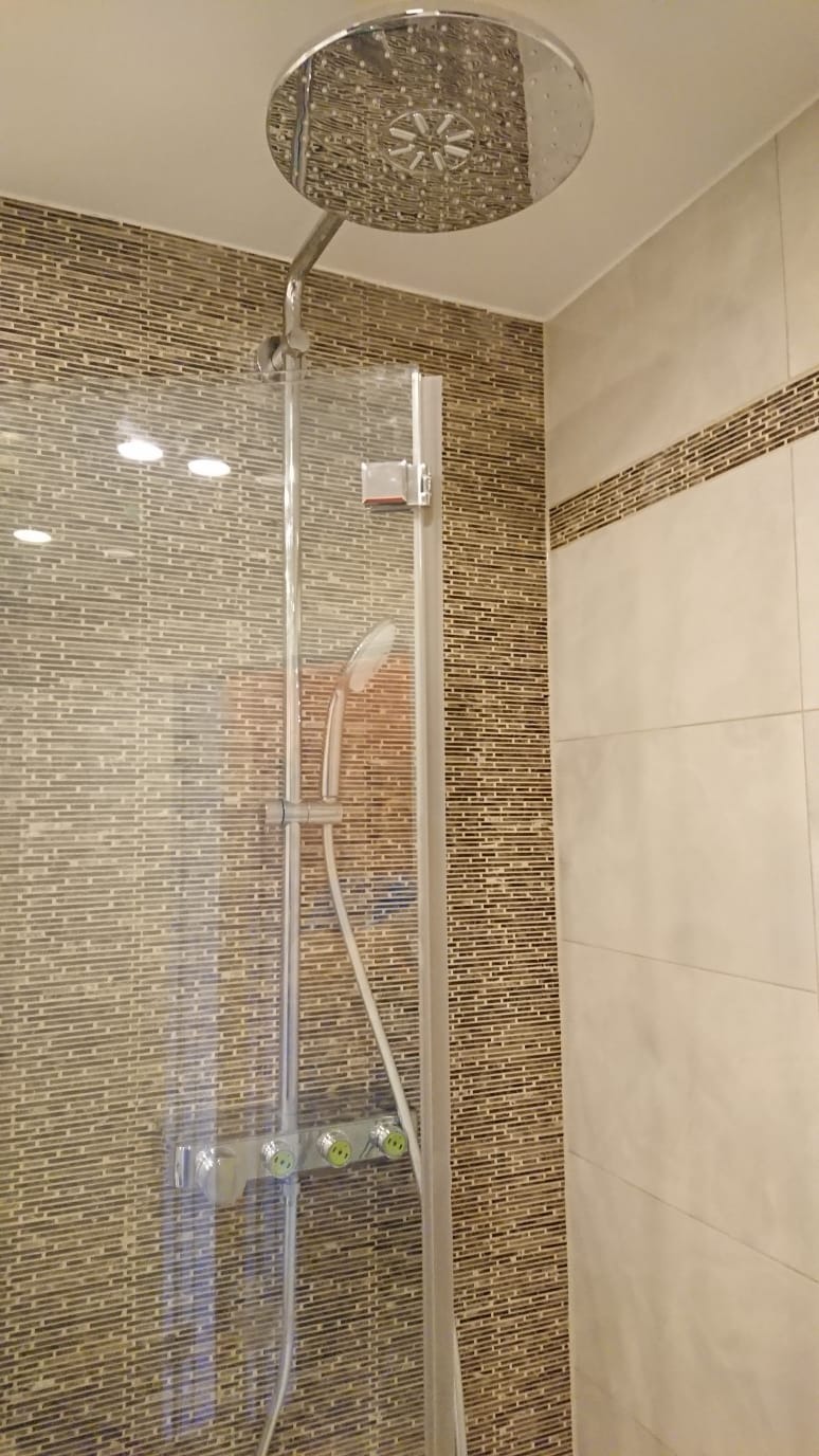 Nowoczesna kabina prysznicowa z deszczownicą i szklaną ścianką, wykończona mozaiką z brązowych płytek i jasnymi panelami ściennymi.