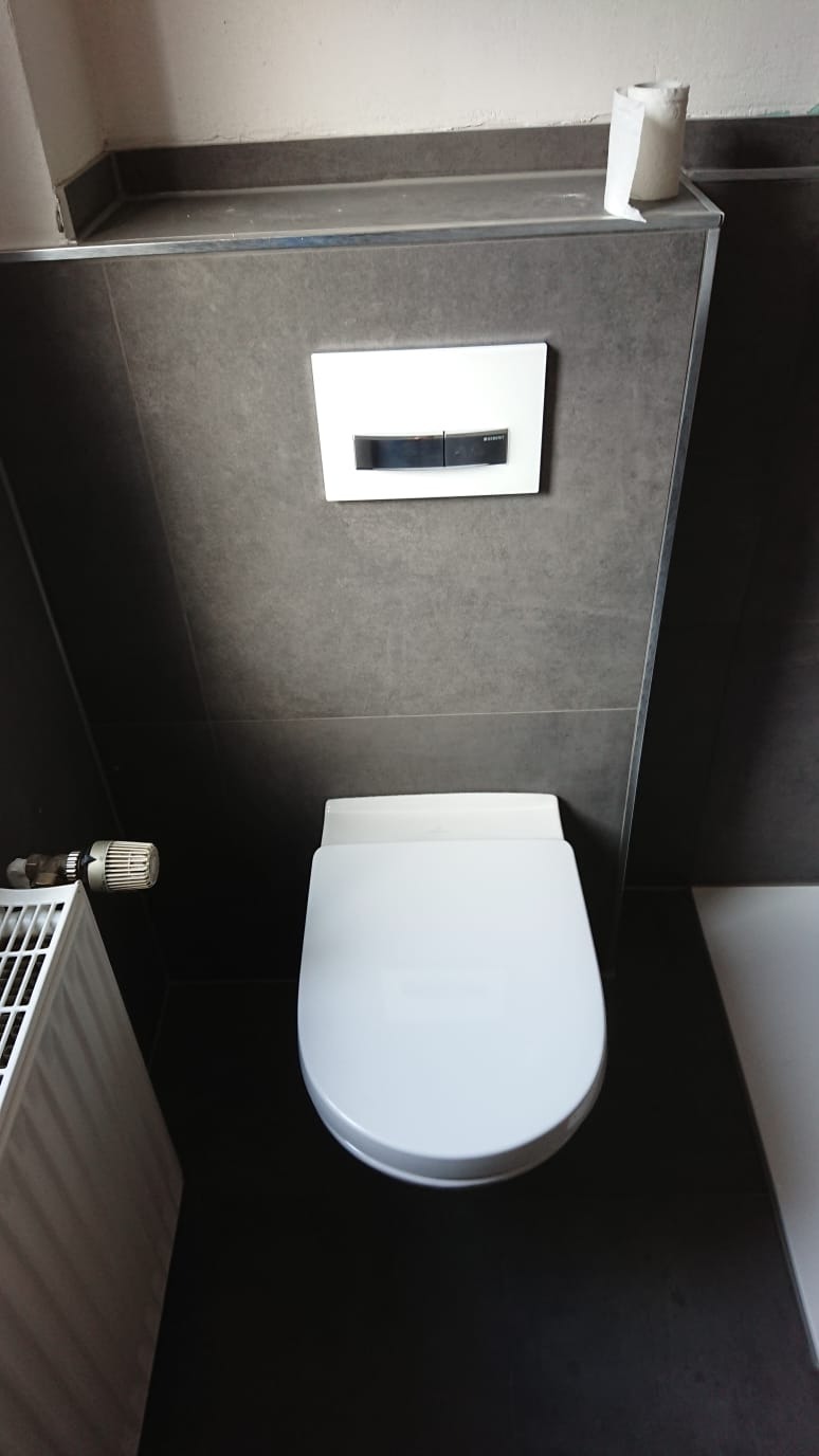 Wisząca toaleta z przyciskiem spłukującym na szarej ścianie, obok grzejnik i rolka papieru toaletowego.