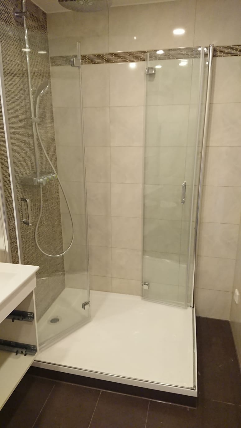 Nowoczesna kabina prysznicowa z częściowo otwartymi szklanymi drzwiami, widoczny zestaw prysznicowy i biały brodzik na tle beżowych płytek z dekoracyjnym paskiem.