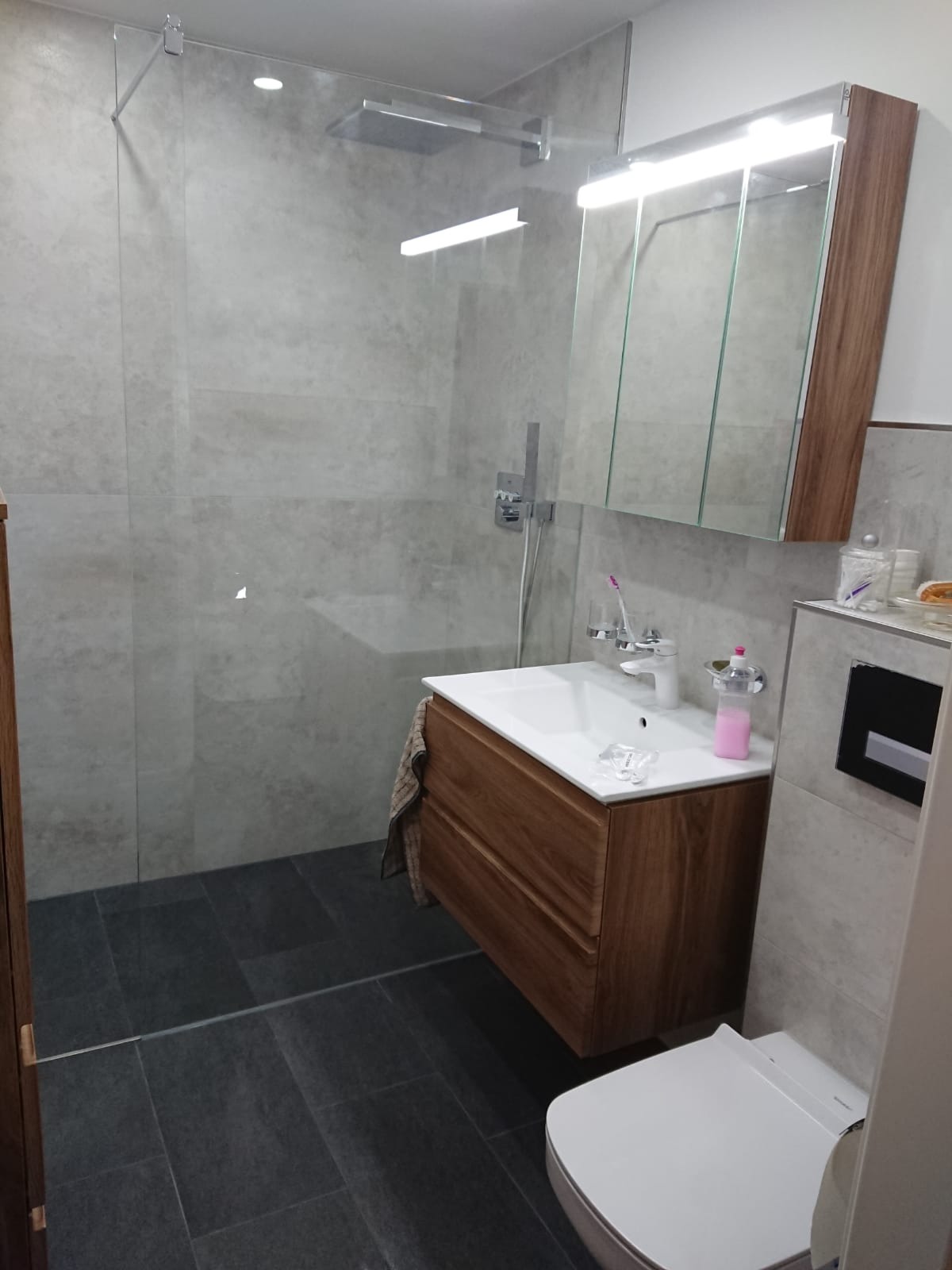 Nowoczesna łazienka z kabiną prysznicową typu walk-in, umywalką nablatową z szafką z drewna, szarą płytką podłogową i wiszącą toaletą.