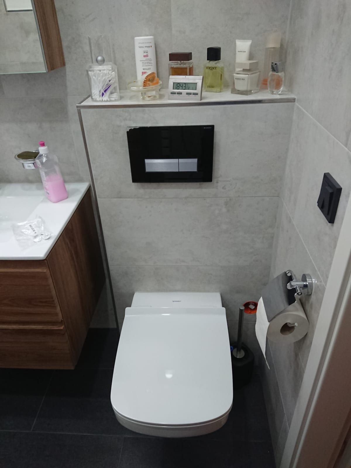 Nowoczesna toaleta wisząca z deską sedesową z funkcją bidetu, czarna spłuczka podtynkowa, szara glazura na ścianach, półka z kosmetykami i akcesoriami łazienkowymi, ciemna podłoga.