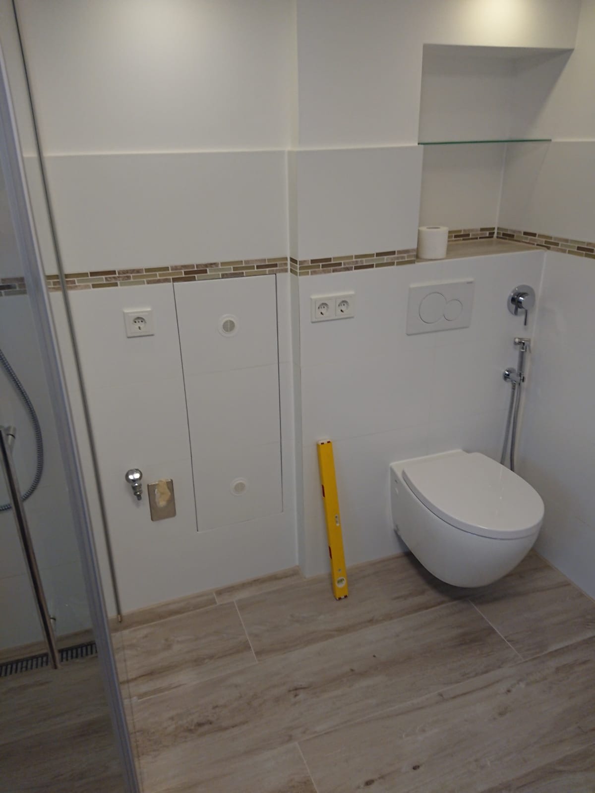 Nowoczesna łazienka z wiszącą toaletą, wykończona białymi płytkami z dekoracyjnym paskiem mozaiki, widoczny poziomica przy ścianie