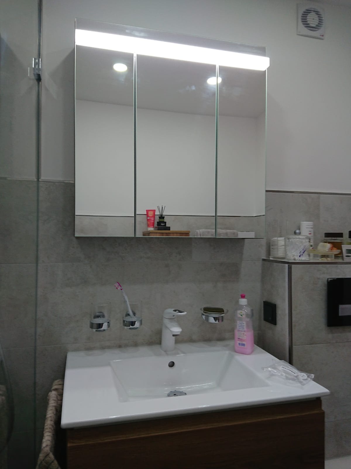 Nowoczesna łazienka z umywalką wpuszczoną w drewnianą szafkę, baterią umywalkową, szarymi płytkami na ścianach i lustrem z oświetleniem LED, na półce kosmetyki i dyfuzor zapachowy.