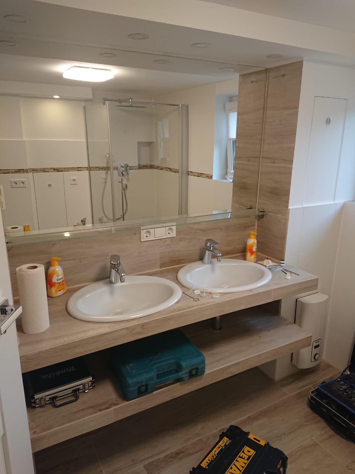 Dwie białe umywalki nablatowe z chromowanymi bateriami w jasnej łazience, widoczne narzędzia i skrzynki narzędziowe sugerujące trwający remont, duże lustro odbija kabinę prysznicową.