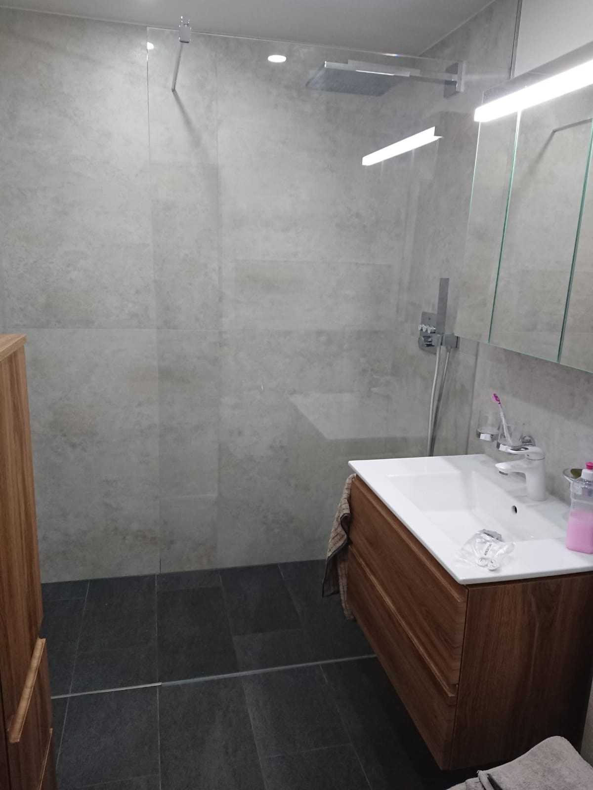 Nowoczesna łazienka z kabiną prysznicową typu walk-in, szarymi płytkami imitującymi kamień na ścianach i ciemną podłogą, z drewnianą szafką pod umywalką i minimalistycznym oświetleniem.