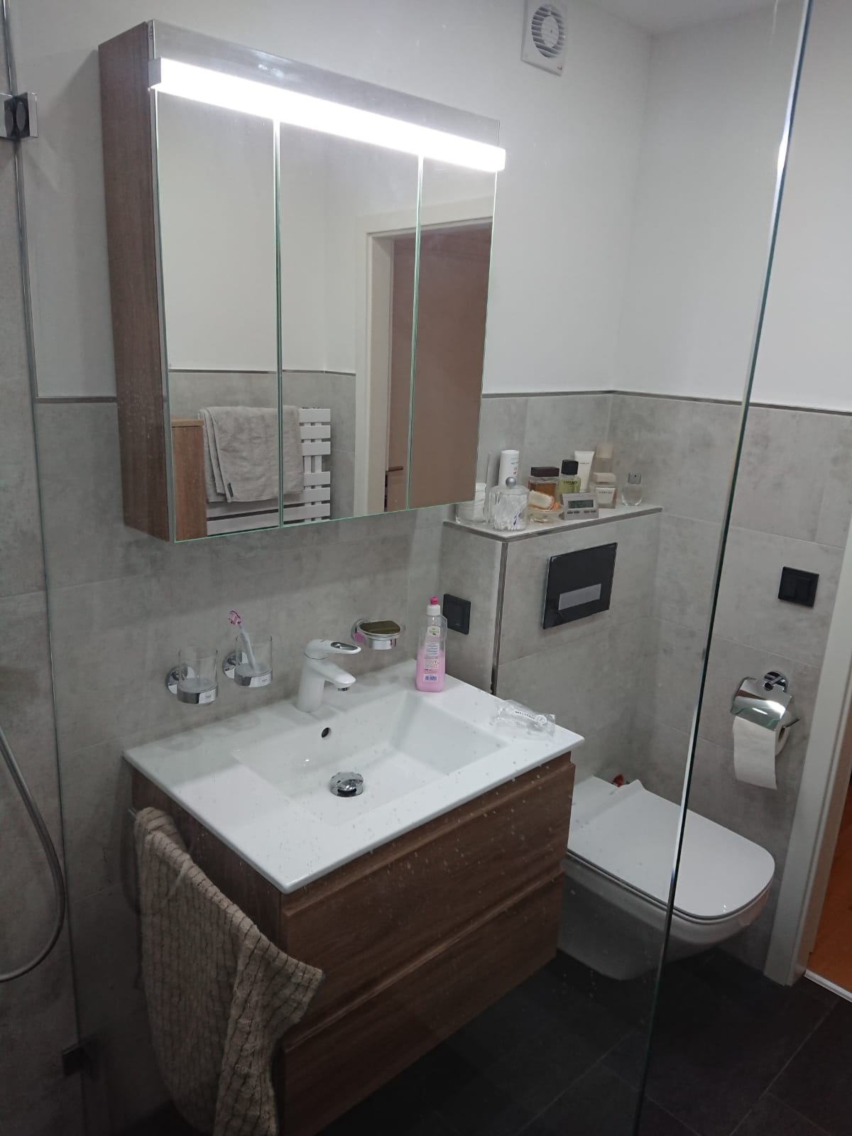 Umywalka z szafką w kolorze drewna, biała ceramika sanitarna, szare płytki na ścianach, lustro z oświetleniem górnym, wentylator sufitowy i szklana kabina prysznicowa w tle.