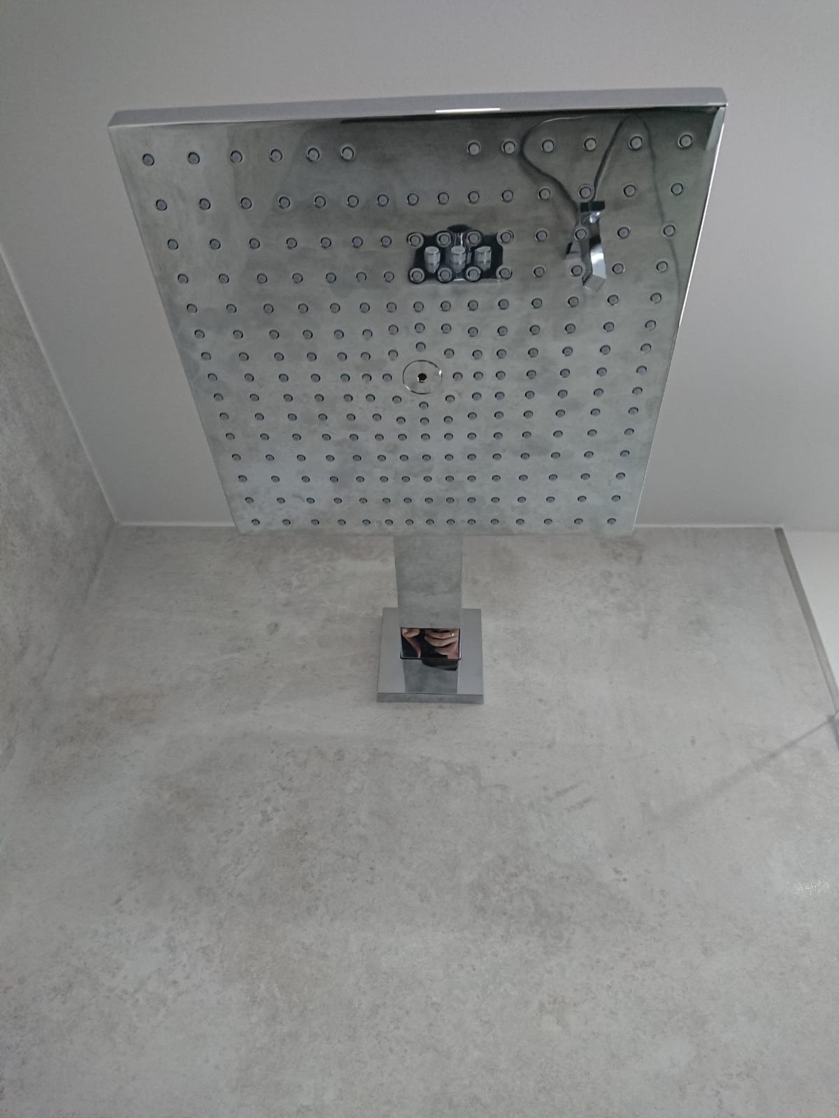 Kwadratowa deszczownica chromowana zamontowana na suficie w łazience z szarą, imitującą beton ścianą.