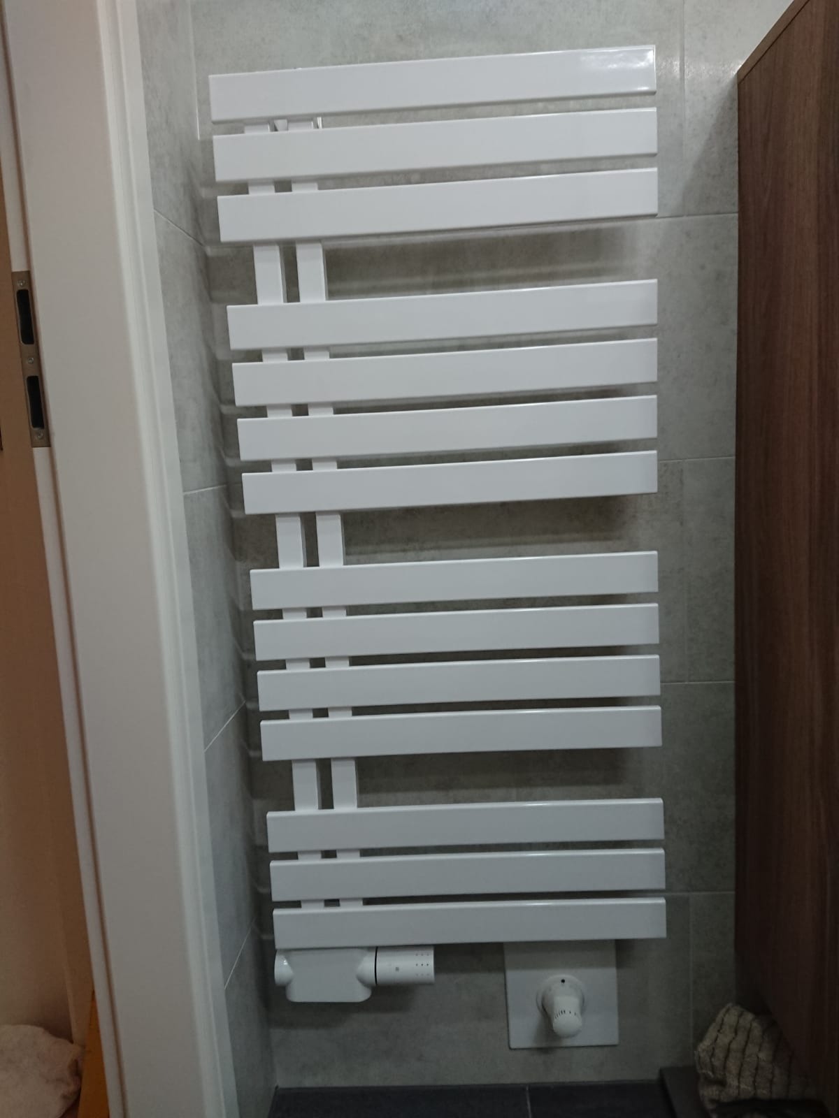 Biały, drabinkowy grzejnik łazienkowy na szarej, kafelkowanej ścianie, z widocznym termostatem i elementami instalacji.