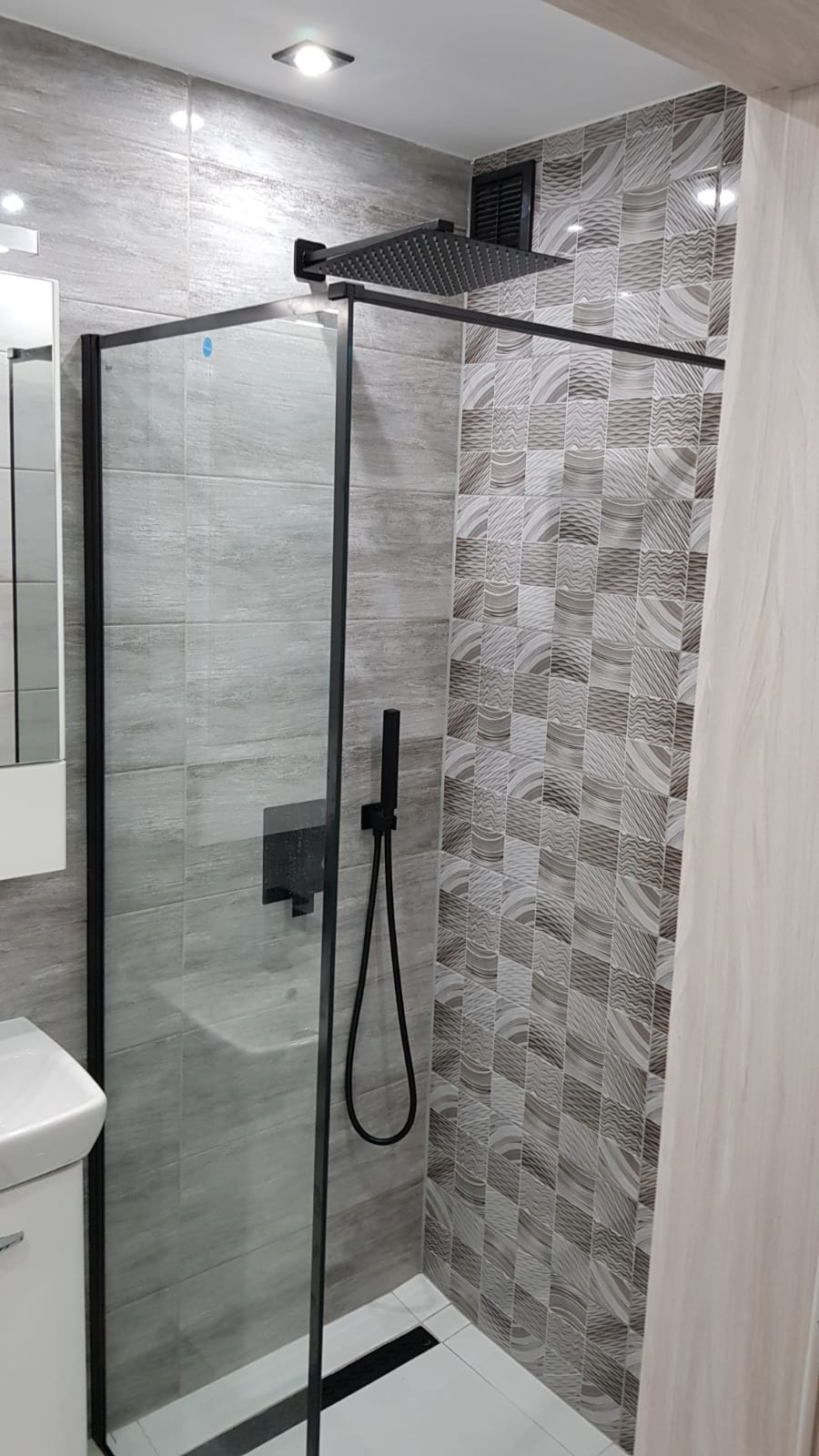 Nowoczesna kabina prysznicowa z czarną armaturą i deszczownicą, szare płytki imitujące kamień oraz dekoracyjne płytki mozaikowe w odcieniach szarości i beżu.