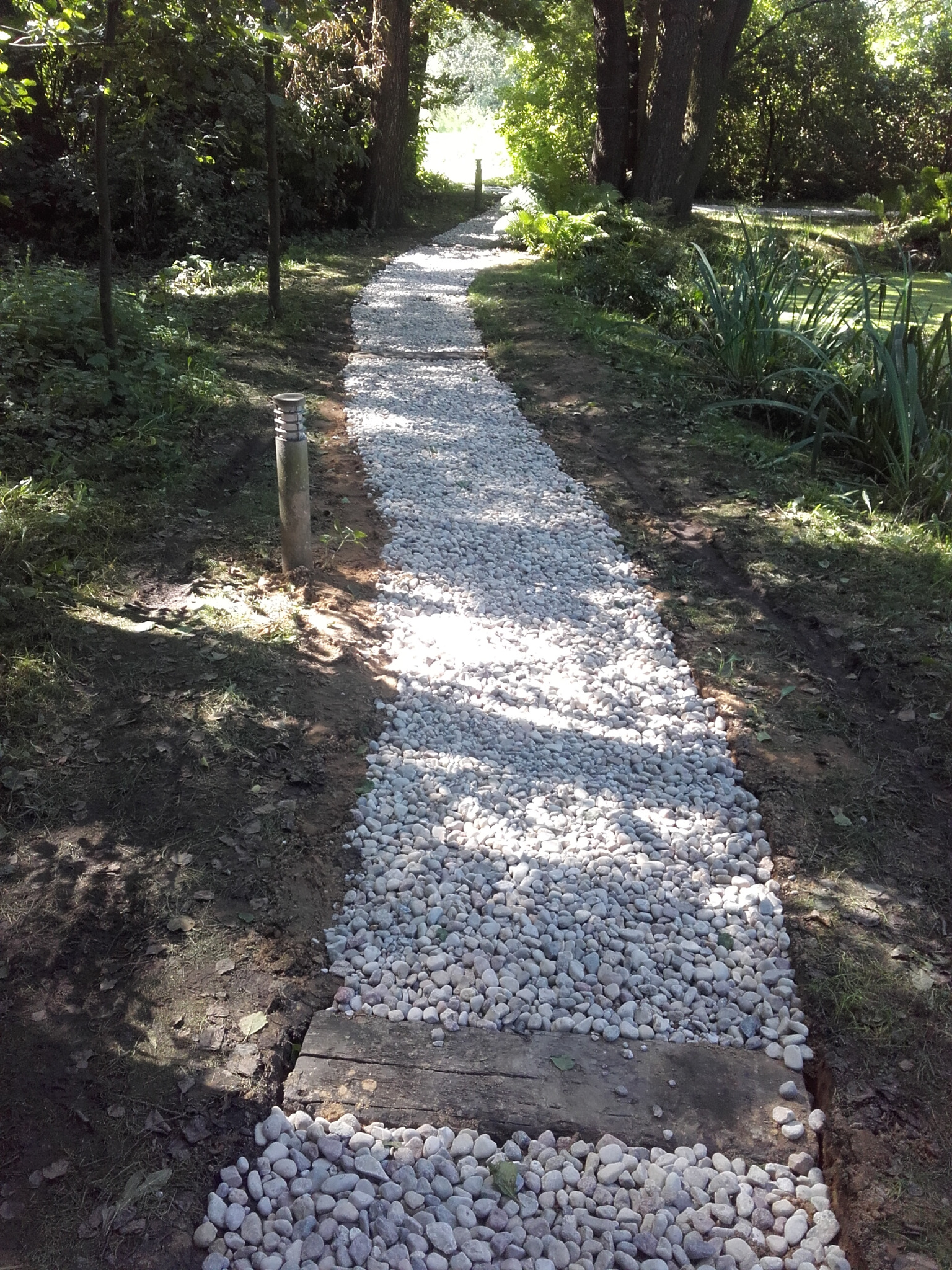 Ścieżka w ogrodzie wysypana białym żwirem, obramowana drewnianymi belkami, prowadząca przez zacieniony teren w kierunku jasnego punktu w oddali.