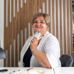 Kobieta w białym fartuchu prezentuje wachlarz wzorów zdobień paznokci, siedząc za białym stołem w salonie kosmetycznym z drewnianą ścianą w tle.