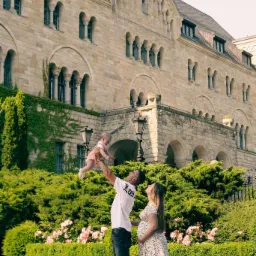 Sesja ciążowa: mężczyzna podnosi niemowlę na tle monumentalnego, kamiennego budynku z licznymi oknami łukowymi, a obok stoi uśmiechnięta, ciężarna kobieta w kwiecistej sukience.