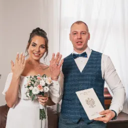 Para młoda prezentuje obrączki i akt ślubu z Urzędu Stanu Cywilnego, ona trzyma bukiet z białych i różowych róż, on ma na sobie niebieską muchę i kamizelkę w kratę.