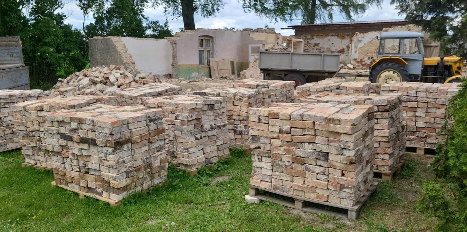 Stosy odzyskanej cegły na paletach, pozostałości po rozbiórce budynku, widoczny traktor i przyczepa.